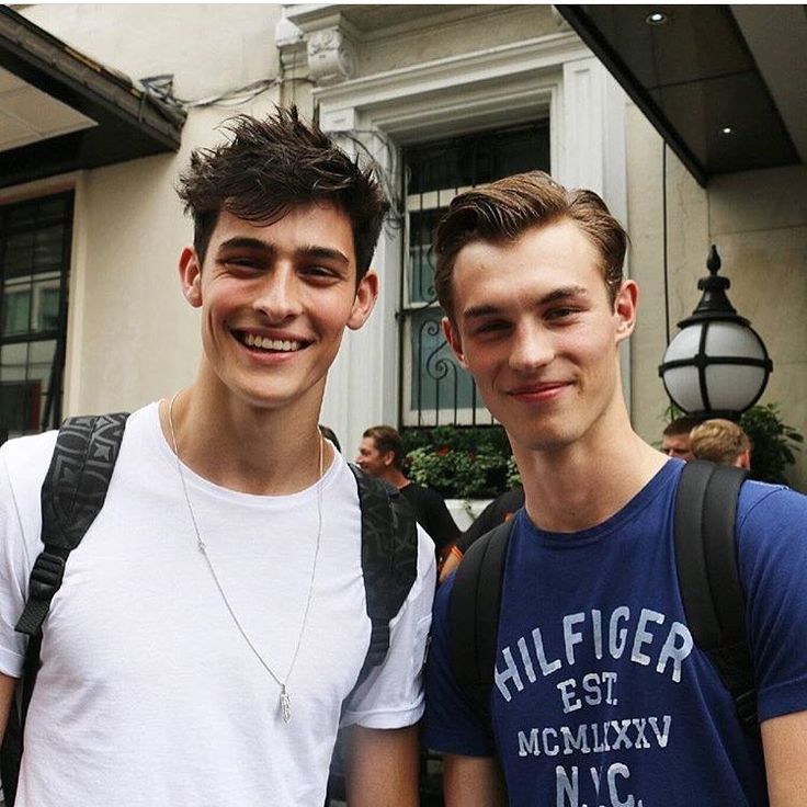 two young men standing next to each other in front of a building with people walking by