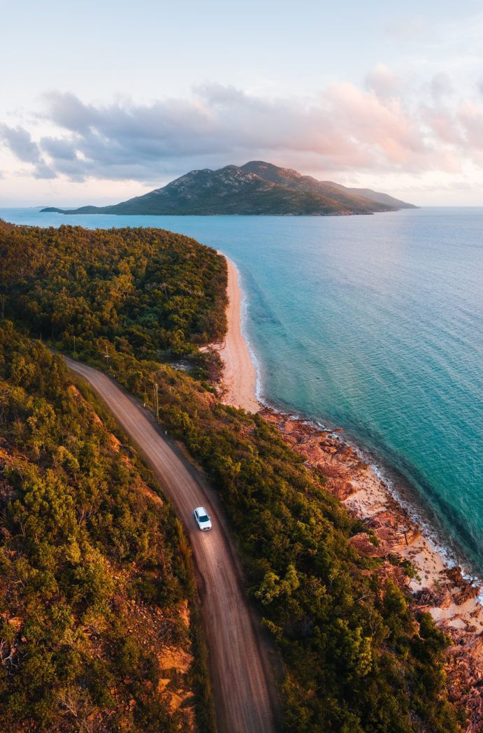 an aerial view of a car driving down a road near the beach and ocean with mountains in the background