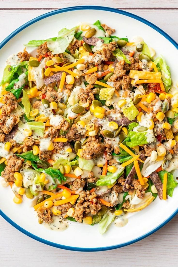 a white plate topped with lettuce, corn and ground beef salad on top of a wooden table