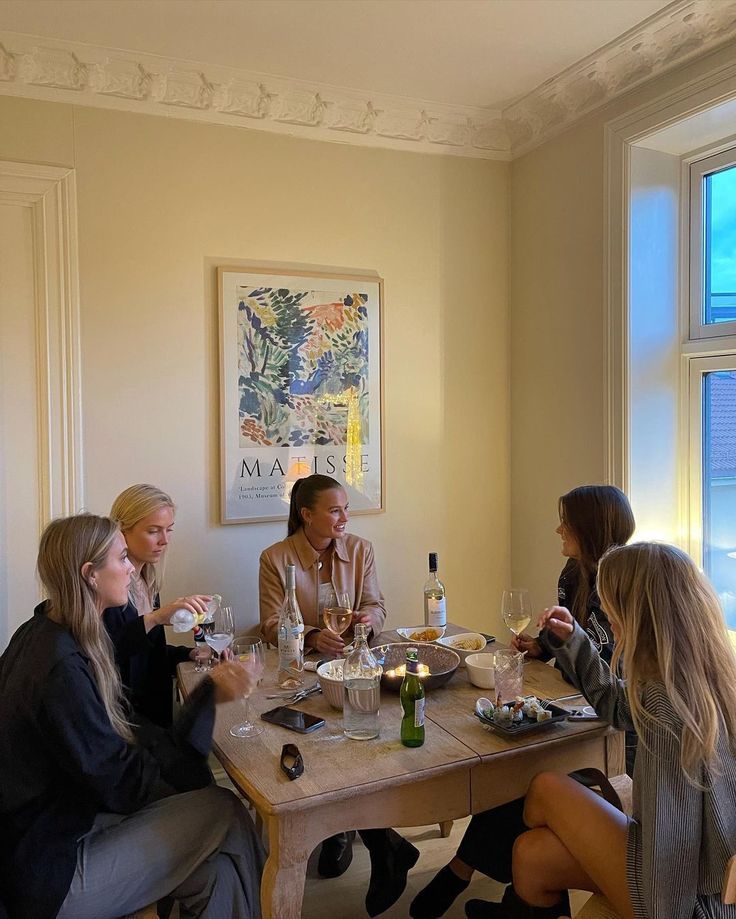 a group of women sitting around a wooden table eating food and drinking wine in front of a window