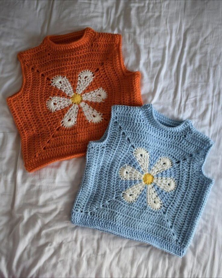 two crocheted sweaters on a bed with white and orange flowers in the middle