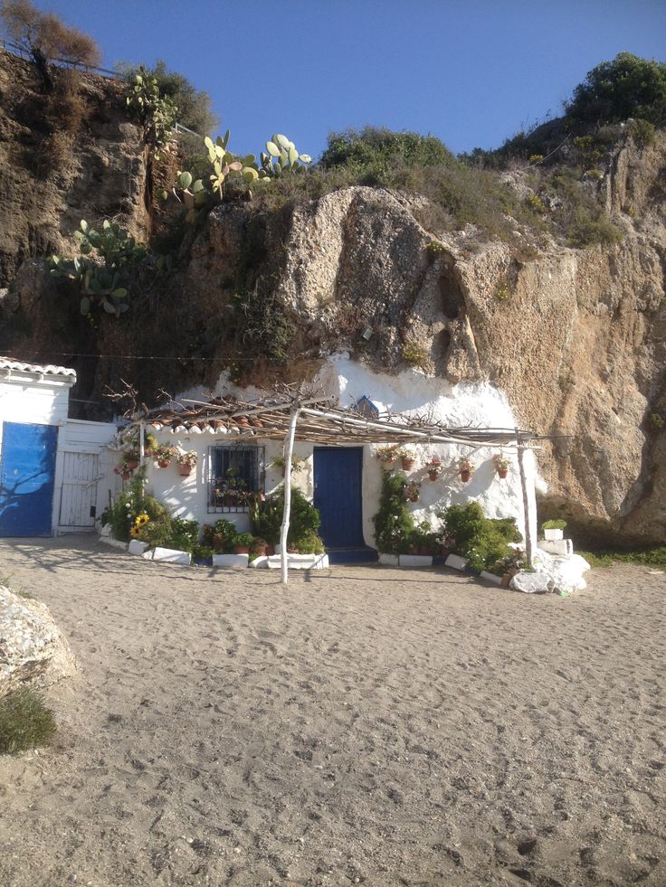 a white house with blue doors and windows on the side of a mountain cliff next to a sandy beach