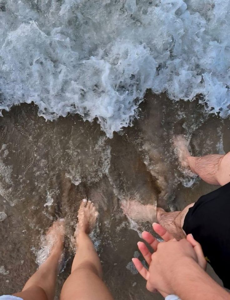 two people are standing on the beach with their feet in the water and one person is holding his hand out