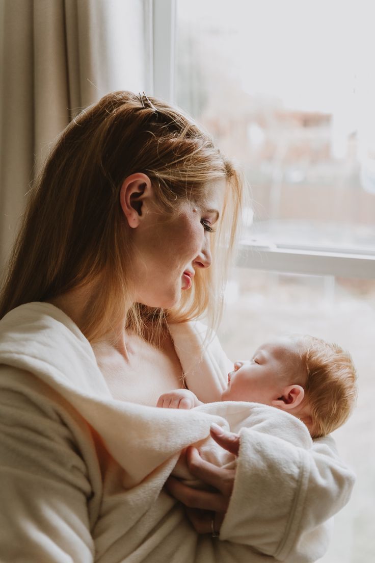 a woman holding a baby wrapped in a blanket while looking out a window at the outside