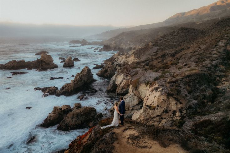 a bride and groom are standing on the edge of a cliff overlooking the ocean with waves crashing against them