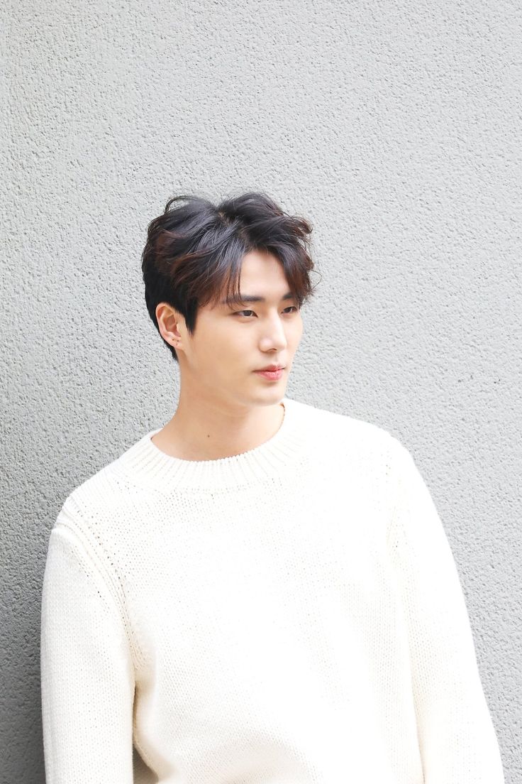 a young man wearing a white sweater standing against a gray wall with his hands in his pockets