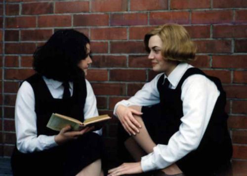 two women sitting next to each other near a brick wall and one is reading a book