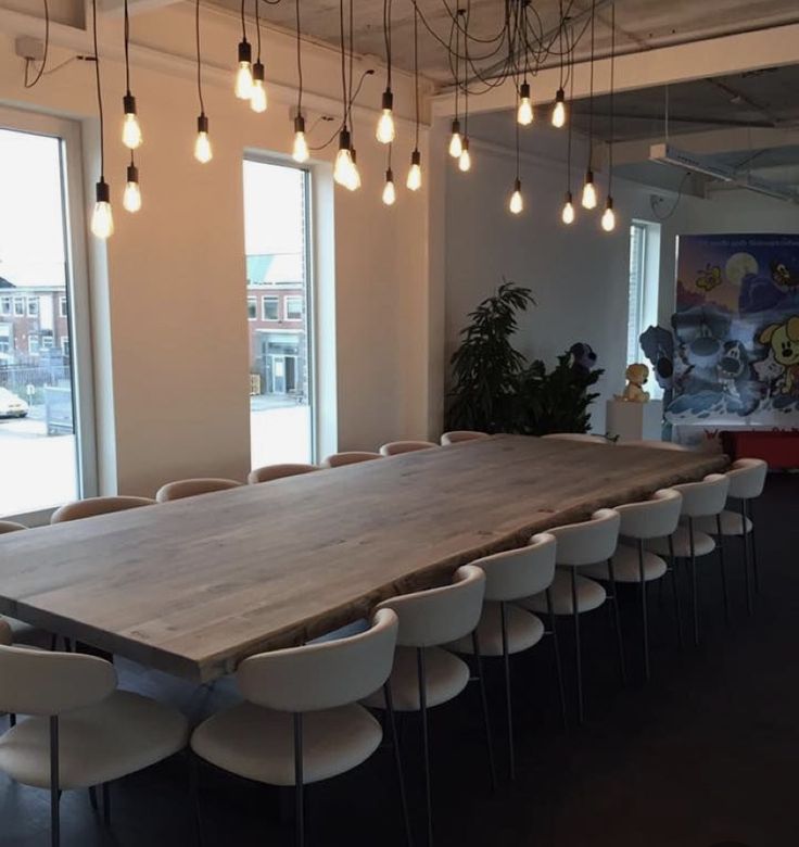 a large wooden table surrounded by white chairs and hanging lights in an office space with windows