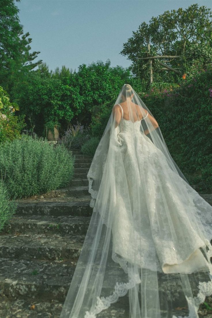 a woman wearing a wedding dress and veil walking up some steps in the grass with her back to the camera