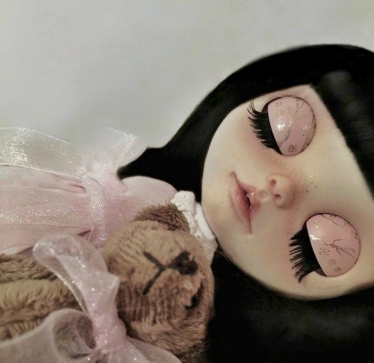 a doll with black hair and pink eyes laying next to a teddy bear