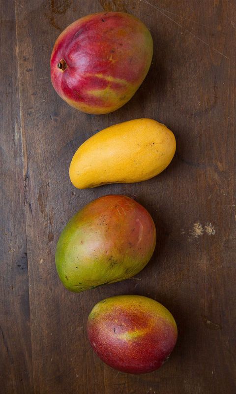 three mangoes, one yellow and one red on a wooden table with other fruit