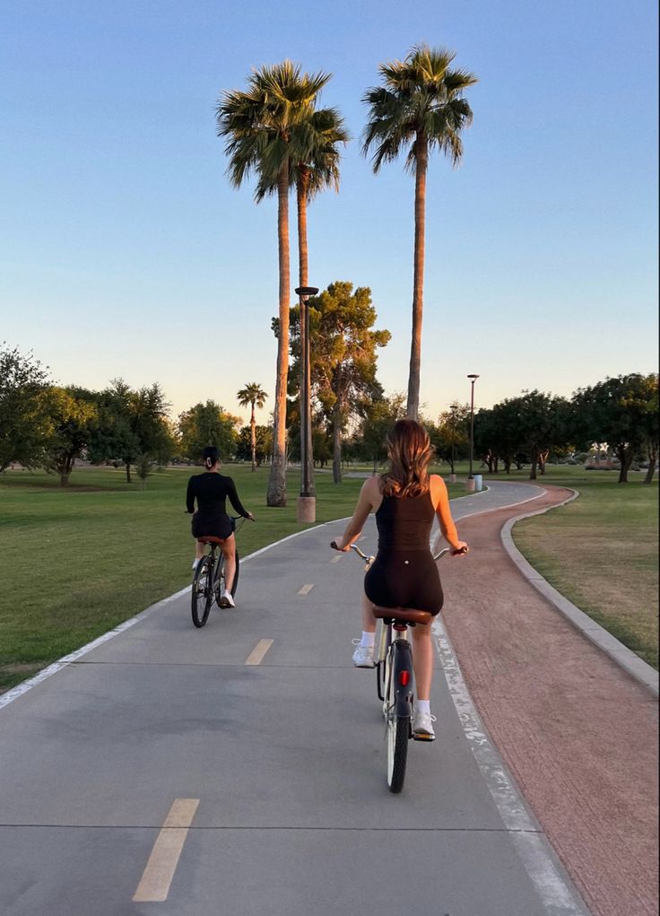 two people riding bikes down a paved path in a park with palm trees and grass