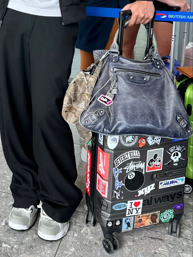 a person standing next to a luggage bag with stickers all over it and holding a handbag