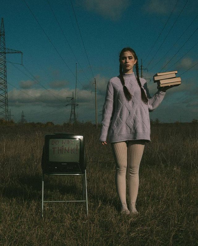 a woman standing in a field holding two books and a television set with wires above her