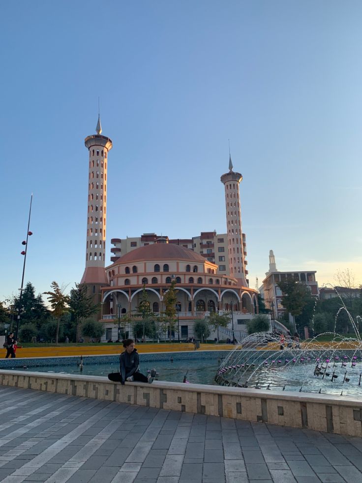 a man sitting on the edge of a fountain in front of a large building with towers