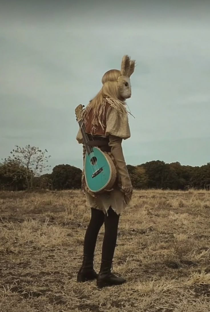 a person in an animal costume with a guitar standing on dry grass and looking off into the distance