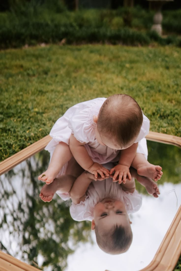 a baby is sitting on top of a mirror in the grass and looking at it's reflection