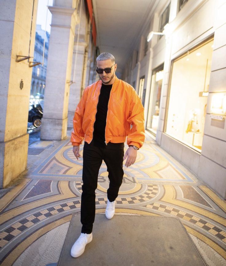 a man in an orange jacket and black pants walking down the street with white shoes