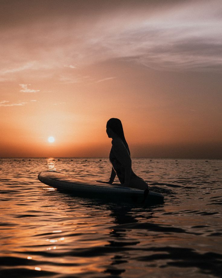 a woman is sitting on a surfboard in the water at sunset or dawn,