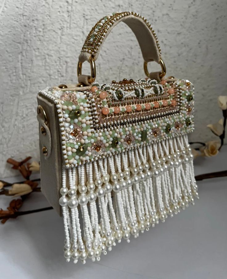 Introducing the Danika Flap Bag, a stunning fusion of bridal elegance and Indian craftsmanship. Hand-beaded with luxurious velvet in enchanting hues of mint, white, and peach, with a beautiful pearl motif, this bag embodies opulence and tradition. Its intricate design, coupled with delicate white suede accents, makes it a standout accessory for brides and lovers of Indian fashion alike. Whether for a wedding ceremony or a cultural celebration, the Danika Flap Bag adds a touch of regal charm to a Luxury Embellished Cream Bags, Luxury Festive Bags With Pearl Handle, White Hand Embellished Party Bag, Embellished Rectangular Shoulder Bag For Receptions, Traditional Evening Shoulder Bag With Pearl Handle, Embellished Rectangular Shoulder Bag For Reception, Bohemian Embroidered Bags For Reception, White Embellished Clutch Bag, Bohemian Embellished Bags For Formal Occasions