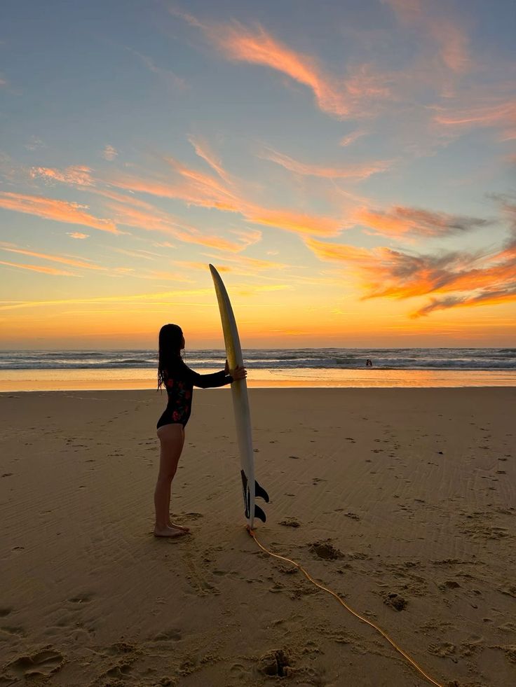 a woman holding a surfboard on top of a sandy beach near the ocean at sunset