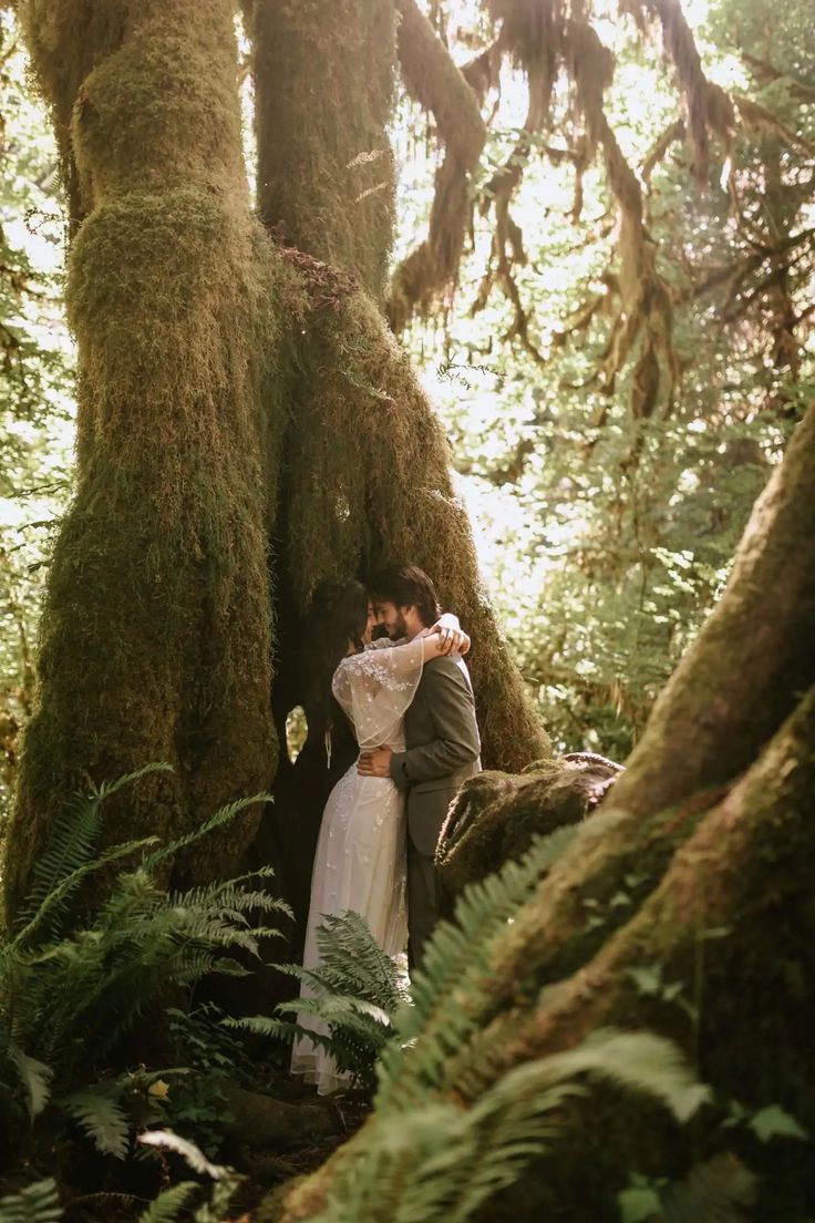 a man and woman standing in the woods next to a large tree with moss growing on it