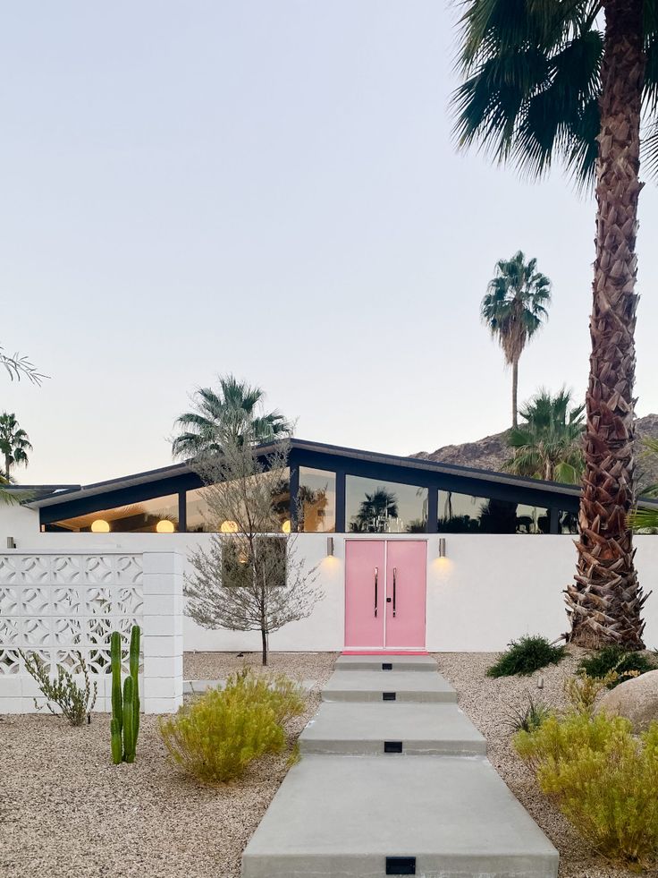 a pink door is on the side of a white house with palm trees around it