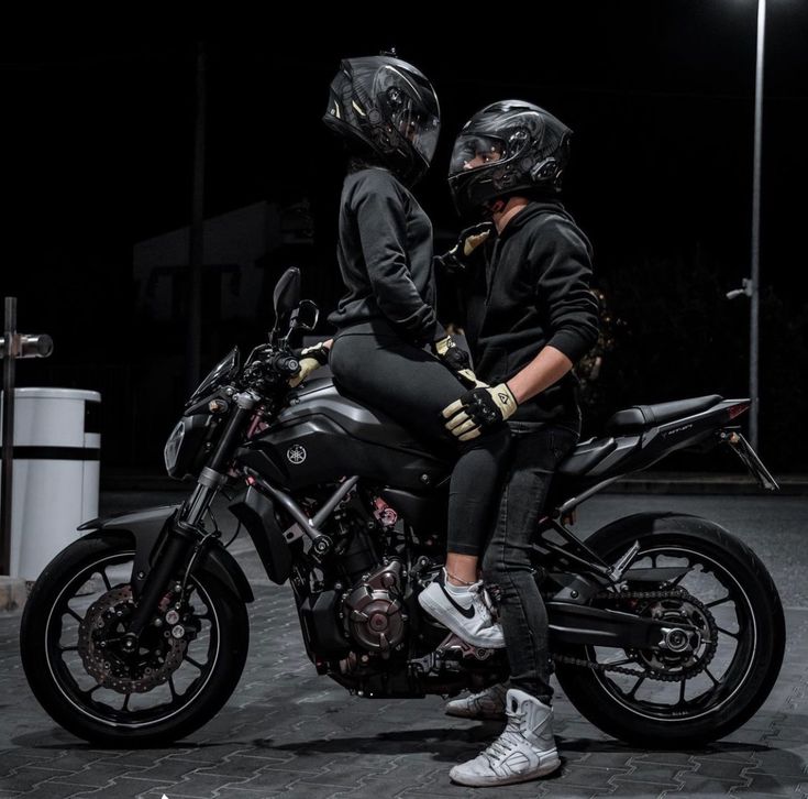 two people sitting on a motorcycle in the dark with their helmets on and looking at something off to the side