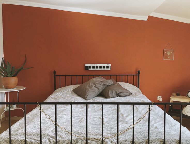 a bedroom with an orange wall and metal bed frame in the foreground is a small table, chair, and lamp on the other side