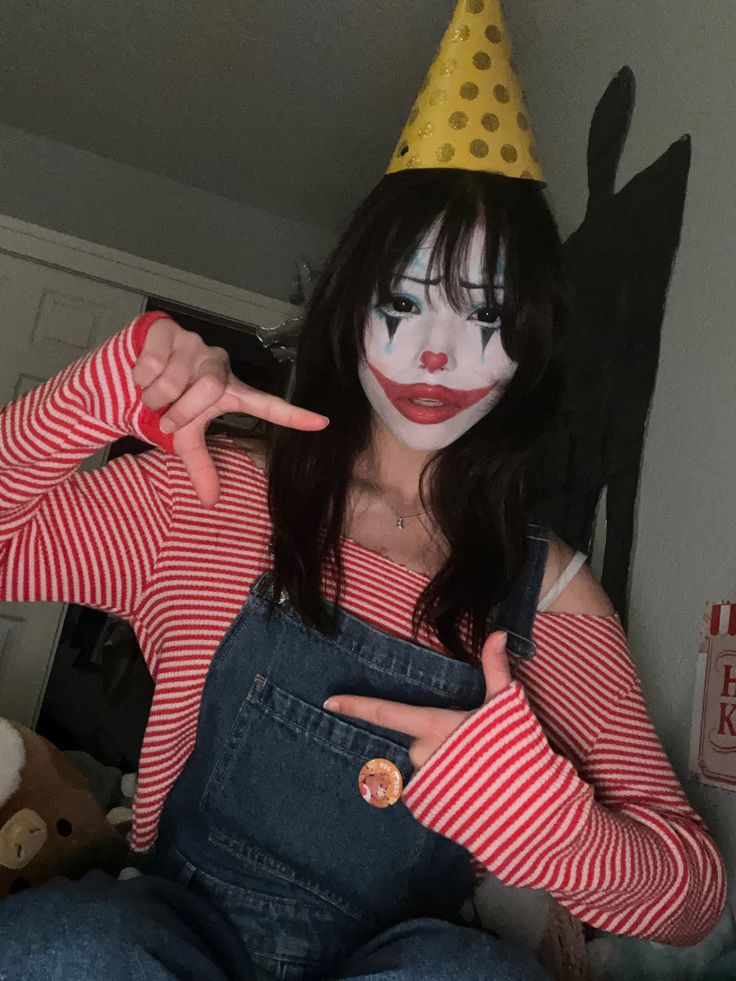 a woman in clown makeup holding her fingers up to the side while wearing overalls and a party hat