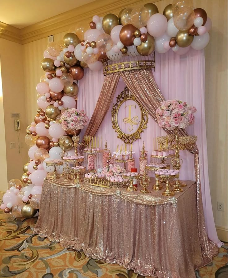 a table topped with lots of pink and gold balloons next to a clock on a wall