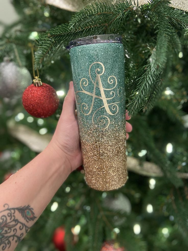 a person holding up a green and gold glitter tumbler next to a christmas tree