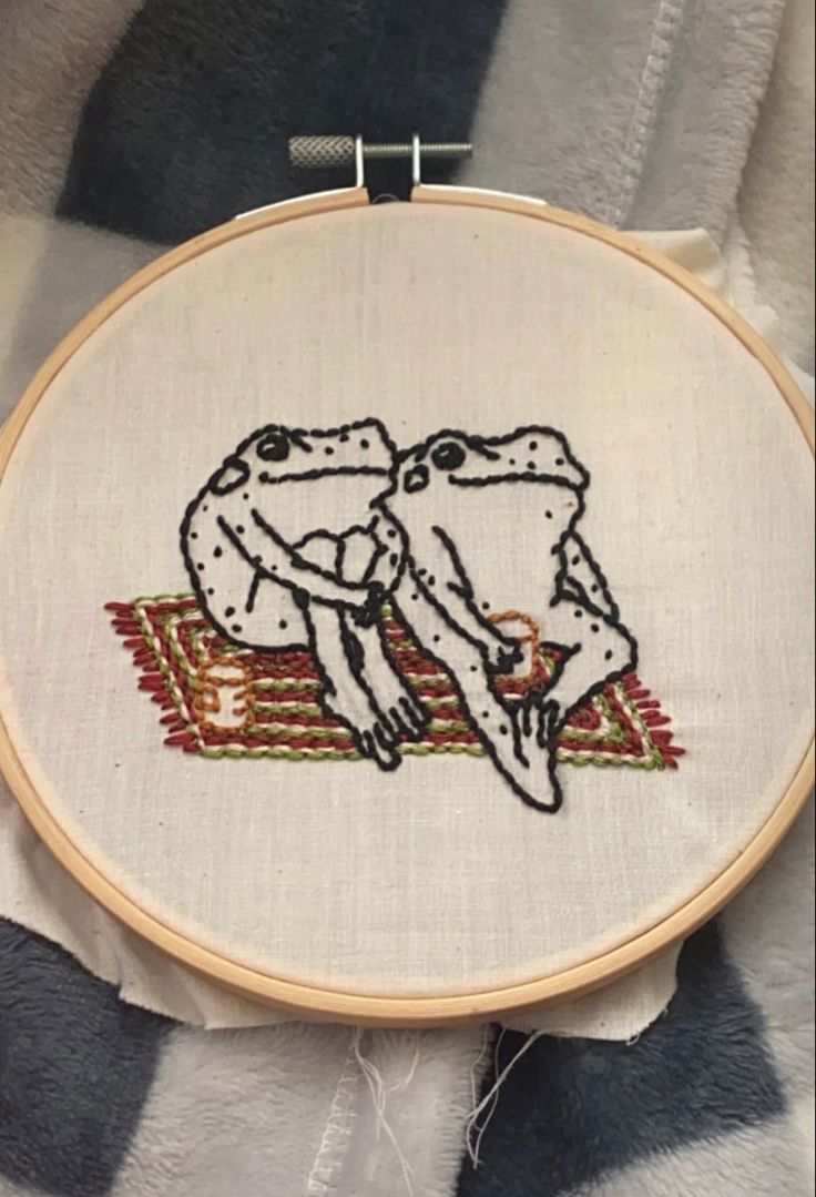 two frogs kissing each other while sitting on a blanket in front of a cross stitch hoop