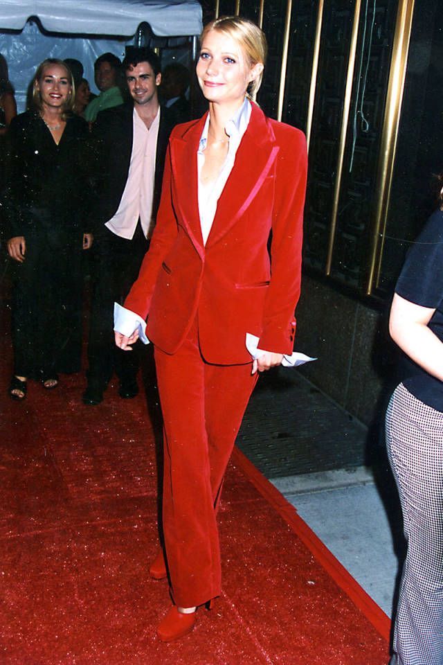 a woman in a red suit and white shirt is standing on a red carpet with other people behind her