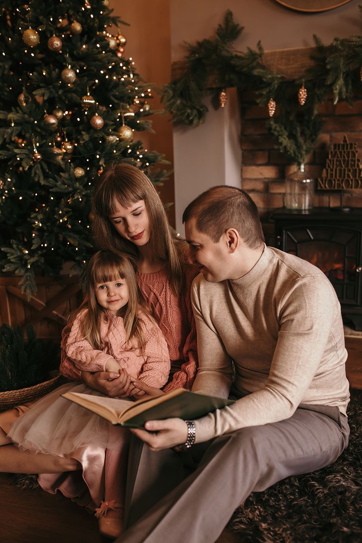 a man, woman and child sitting in front of a christmas tree reading a book