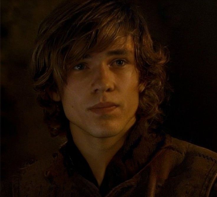 a young man with long hair wearing a brown shirt and looking at the camera while standing in a dark room