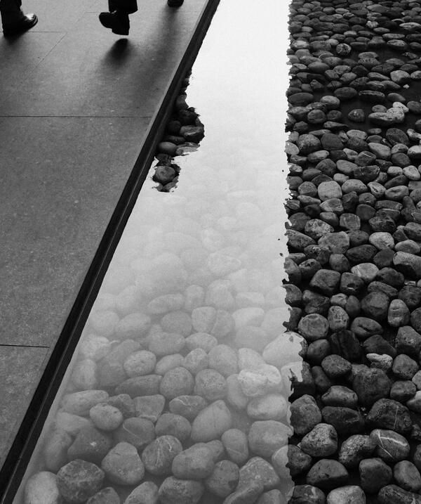 people are walking on the sidewalk near some rocks and water with their reflection in it