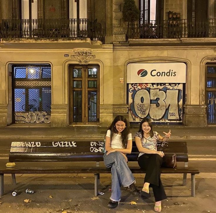 two women sitting on a bench in front of a building with graffiti written on it