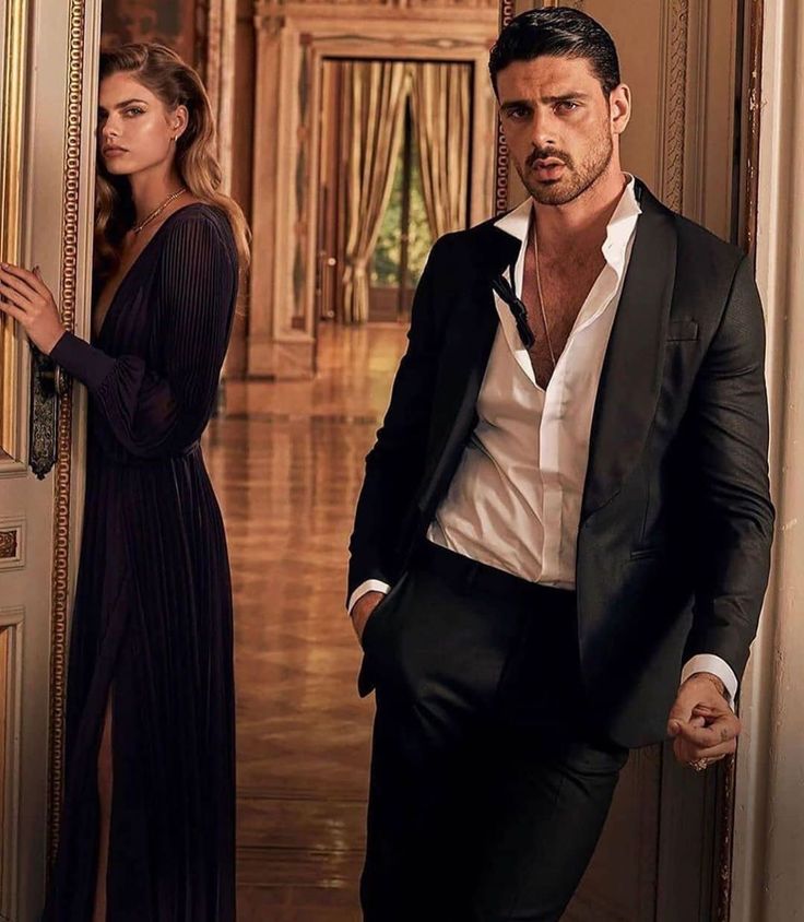 a man standing next to a woman in front of a doorway wearing a black suit and white shirt