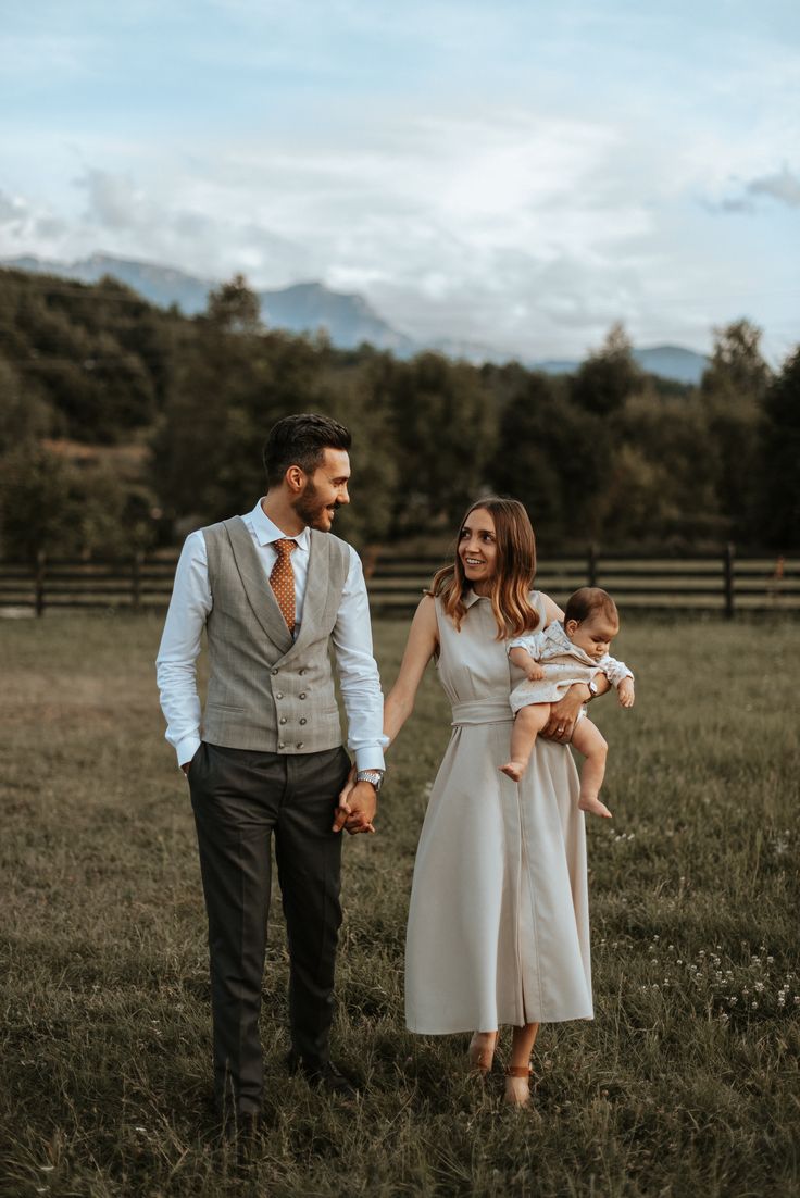 a man and woman holding hands while walking through a field with a baby in their arms