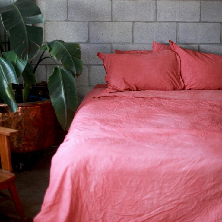 a bed with pink sheets and pillows in front of a brick wall next to a plant