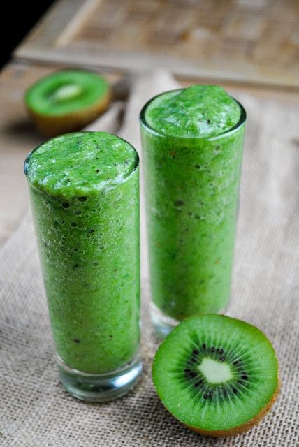 two glasses filled with green smoothie next to an kiwi slice on a cloth