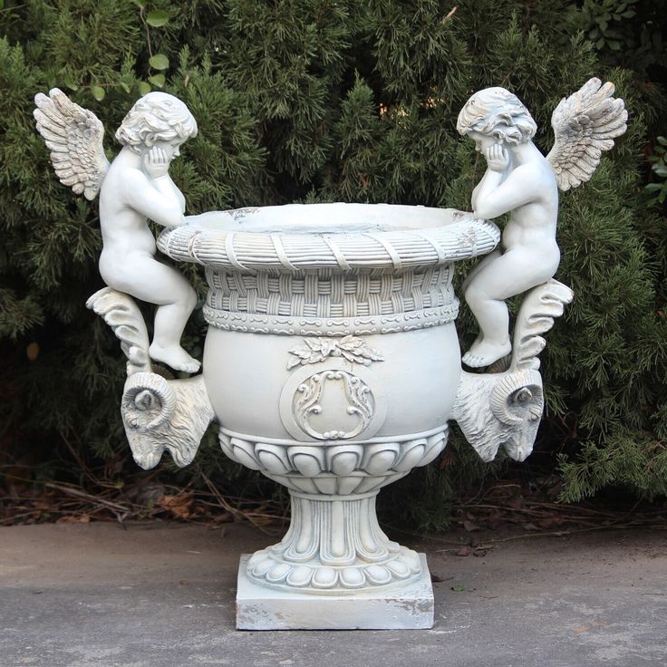 two cherubs sitting on top of a white urn in front of some bushes