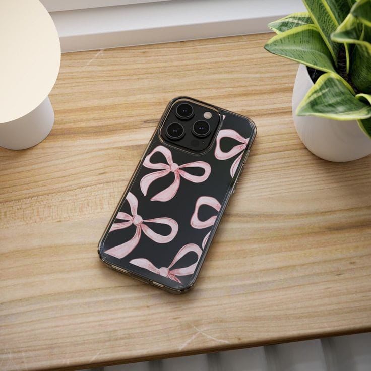 an iphone case with pink bows on it sitting on a wooden table next to a potted plant