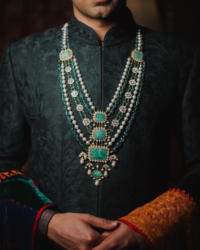 a man wearing a green and gold beaded necklace with pearls on his neck, standing in front of a dark background