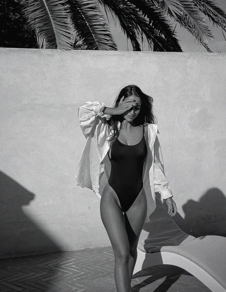 a woman in a swimsuit standing next to a wall and palm tree with her hands on her hips