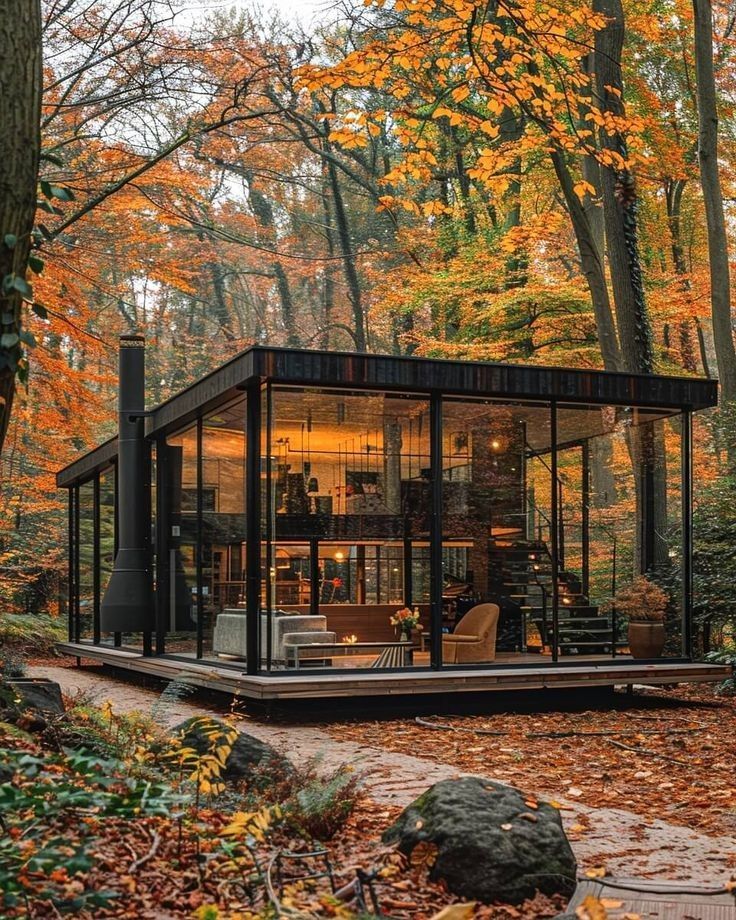 a glass cabin in the woods surrounded by trees with autumn leaves on the ground and foliage