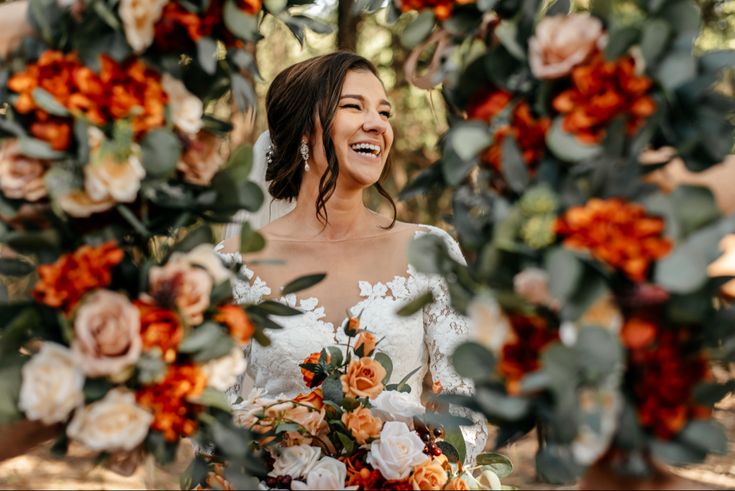 a bride smiles as she stands under an orange and white floral arch with greenery