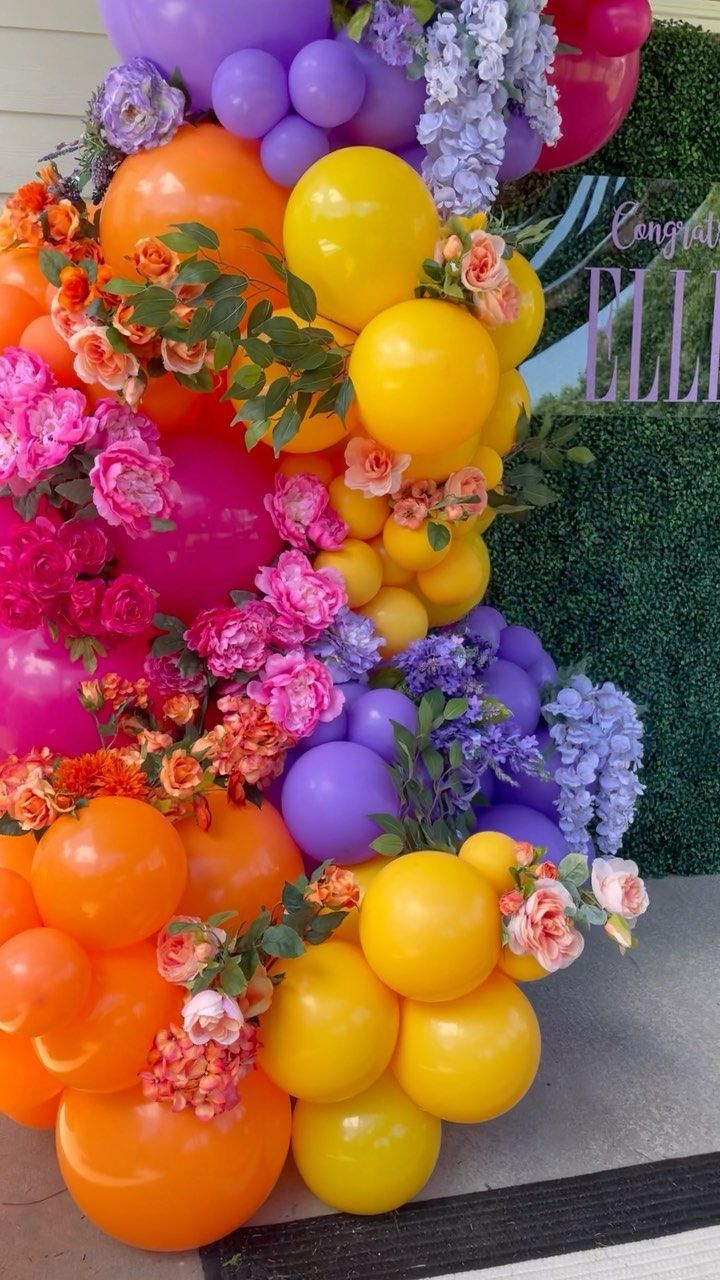 a bunch of balloons that are on the ground next to some flowers and plants in front of a sign