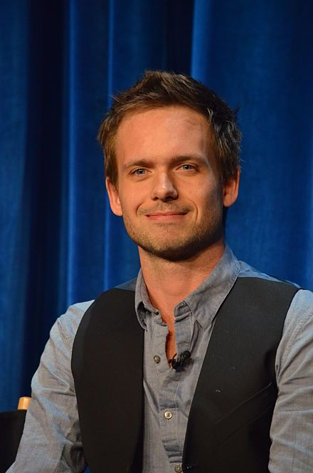 a man in a vest smiles at the camera while sitting next to a blue curtain